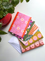 A set of folded note cards with various colorful abstract art designs, displayed with envelopes on a table, with a red potted plant in the background.