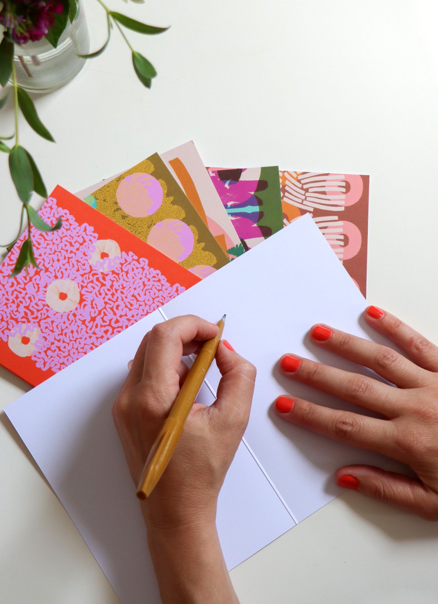 Person writing on a folded note card with colorful cards and a plant in the background