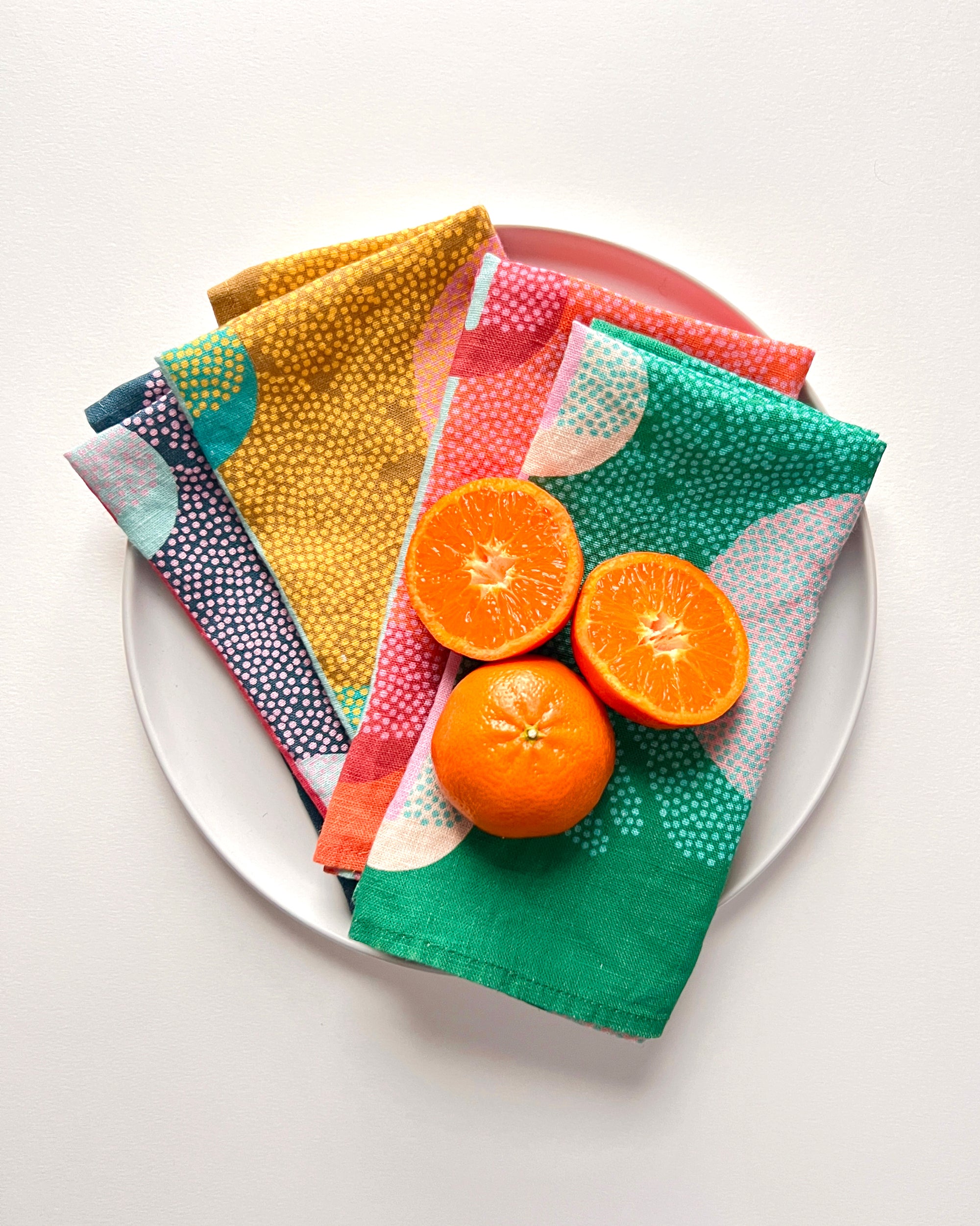 A set of four colorful, unique, artful linen napkins displayed on a plate with a halved orange and several oranges beside the plate.