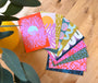 A fan-like spread of colorful postcards with vibrant abstract art patterns, placed on a wooden surface, with a partial view of a plant in the corner.
