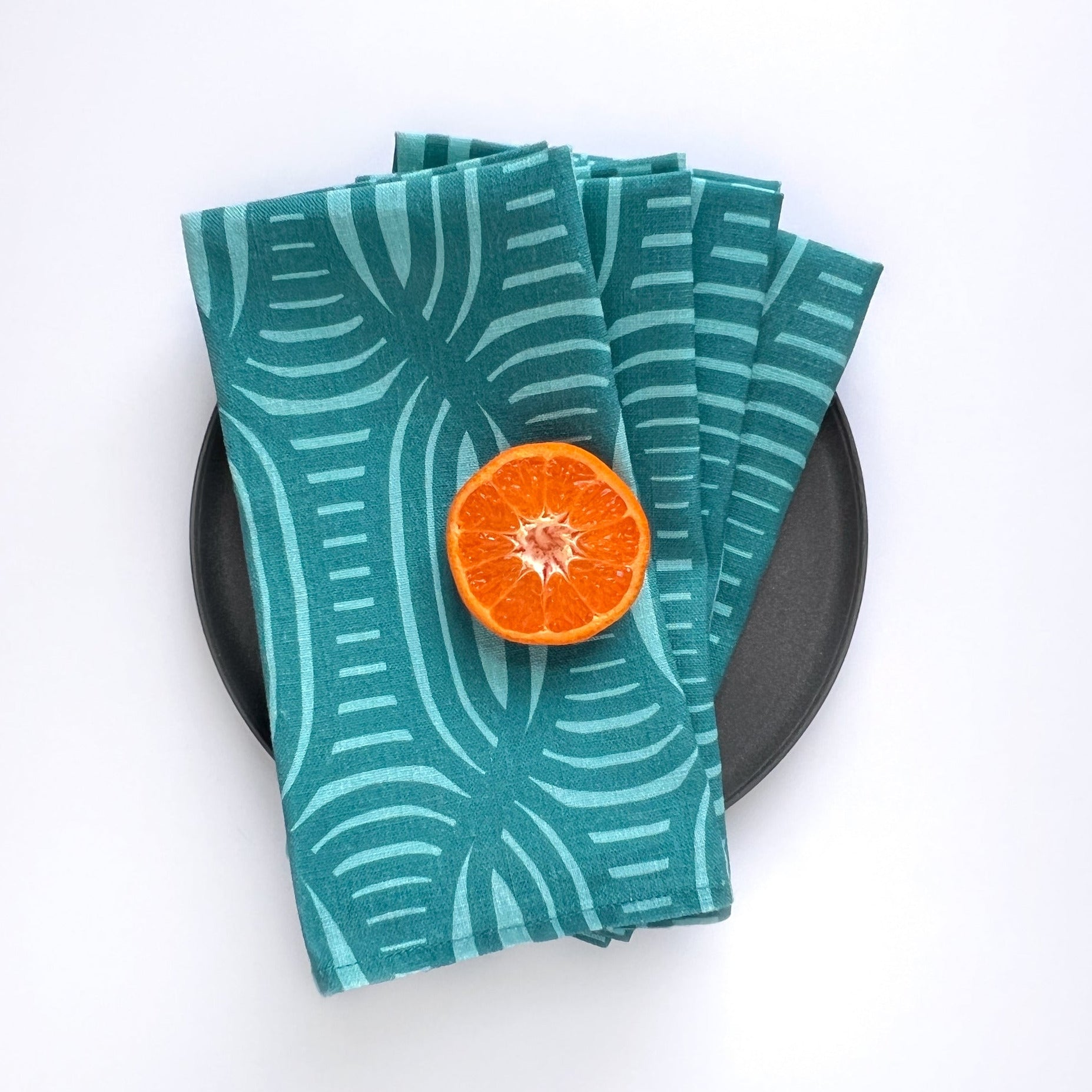 Stack of teal patterned napkins with a halved orange on a black plate against a white background