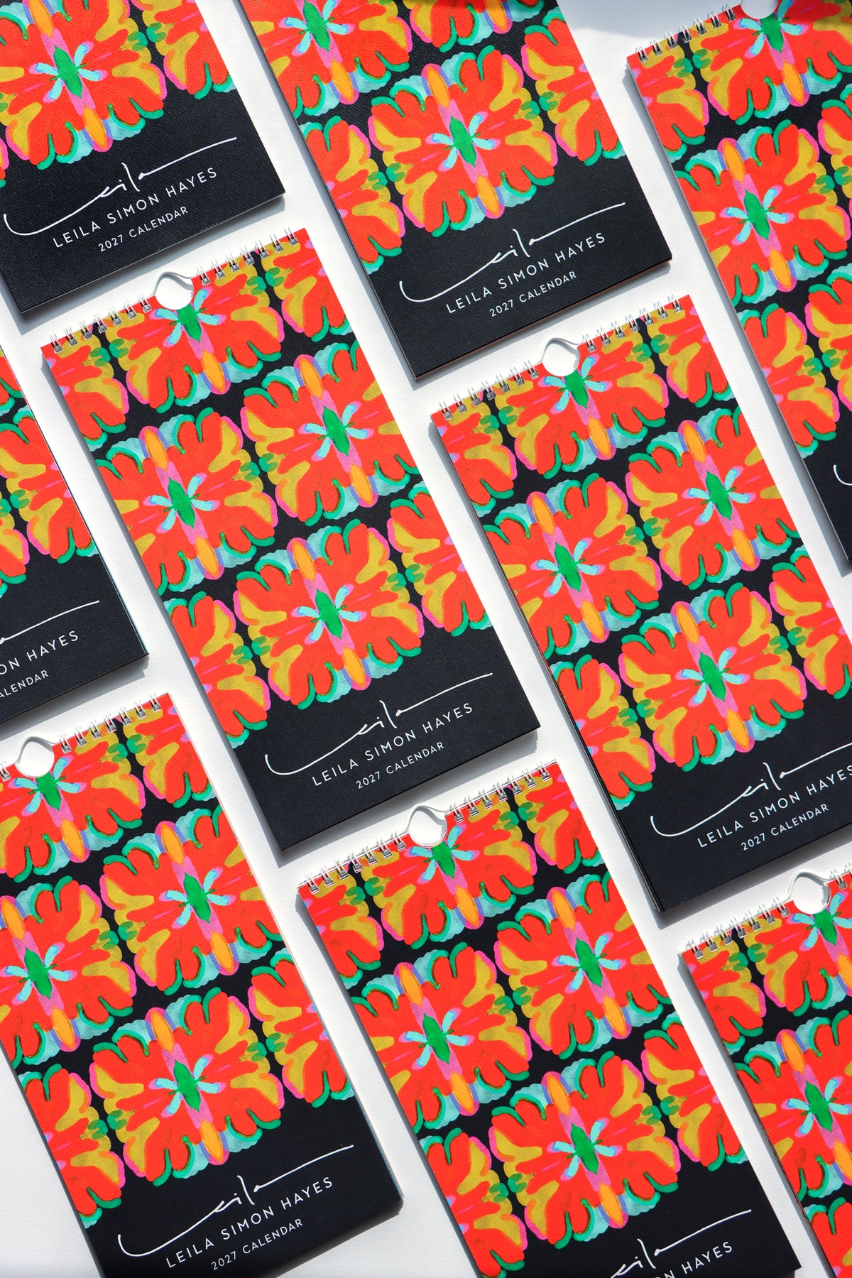 Multiple copies of the Leila Simon Hayes 2021 Calendar arranged in a diagonal grid on a white background. The calendar features a bold, colorful floral pattern with orange, yellow, and green flowers on a black background. Designed by artist Leila Simon Hayes.
