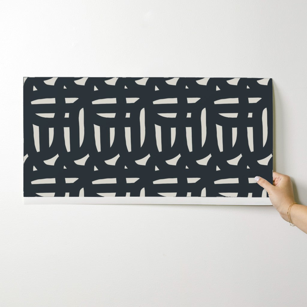 Person holding a rectangular wallpaper swatch with a black and white pattern against a white background. Designed by artist Leila Simon Hayes.