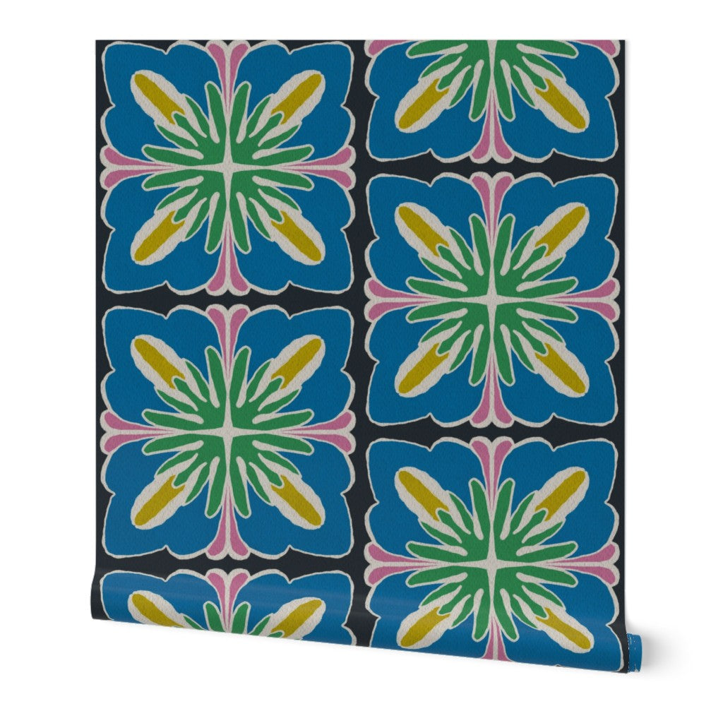 A roll of wallpaper featuring a bold, colorful geometric pattern of six overlapping starburst tiles on a black background. The design includes blue, green, yellow, pink, and white elements, shown against a clean white background. Designed by artist Leila Simon Hayes.