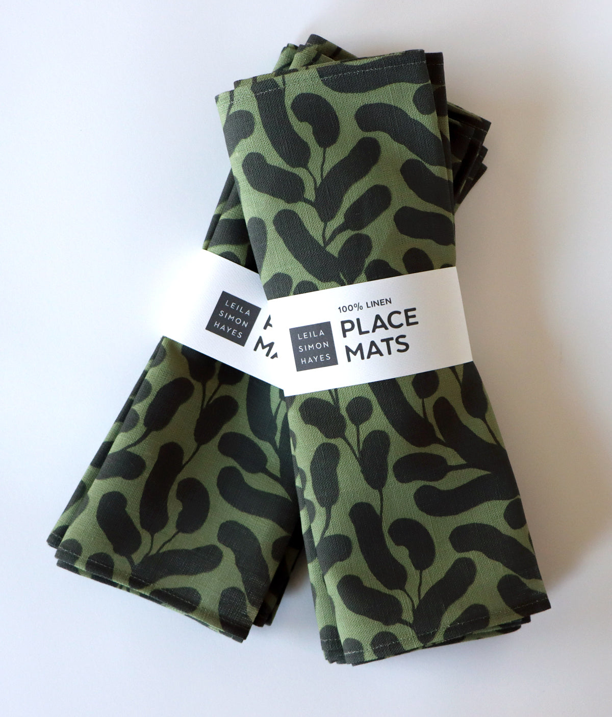 A pack of 4 folded olive green place mats with black leaf patterns wrapped with a label saying "100% linen placemats" designed by artist Leila Simon Hayes on a white background