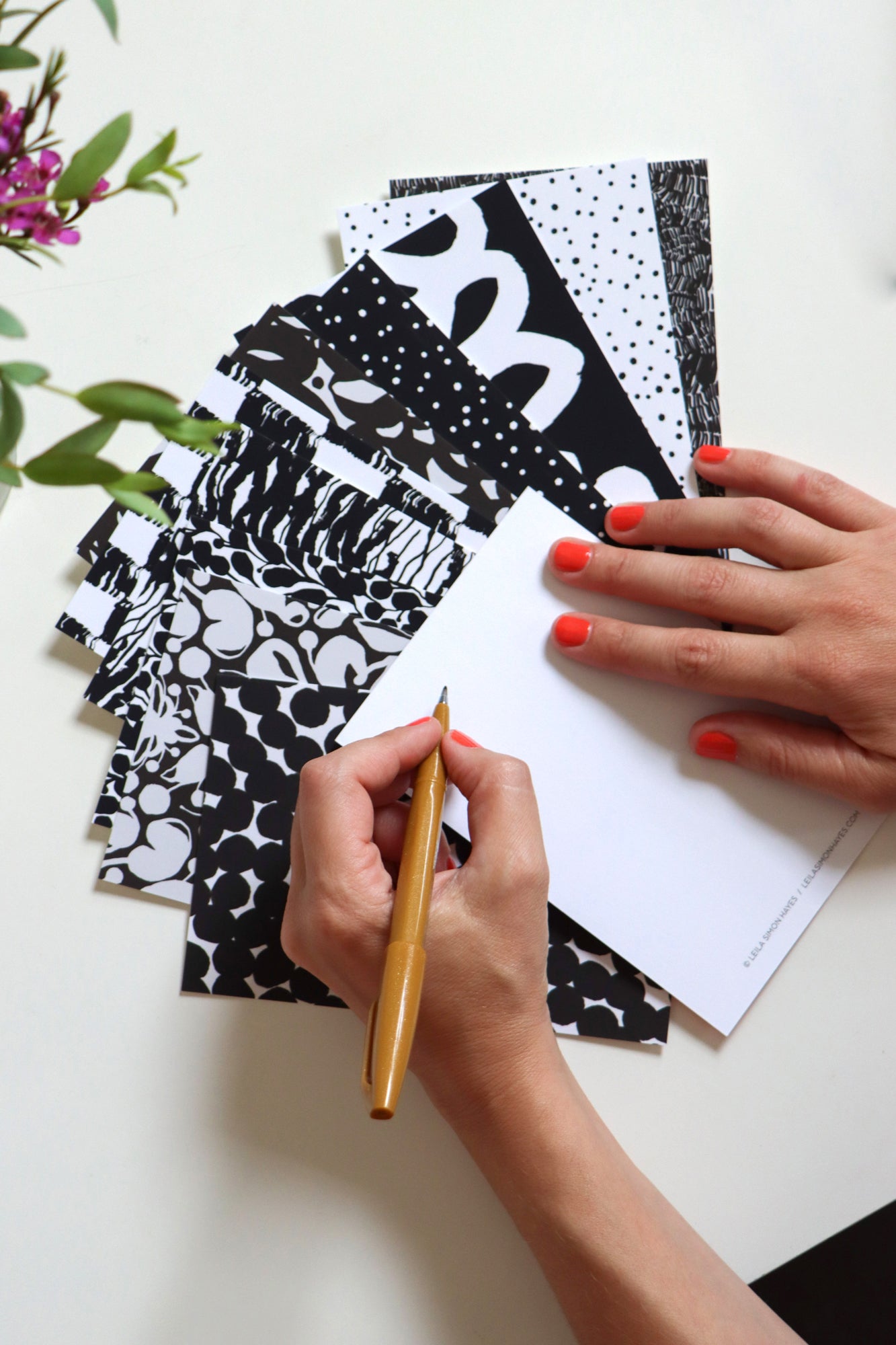 Person holding a pen and writing on the blank side of the postcards with black and white postcards spread across the table in the background. Designed by artist Leila Simon Hayes.