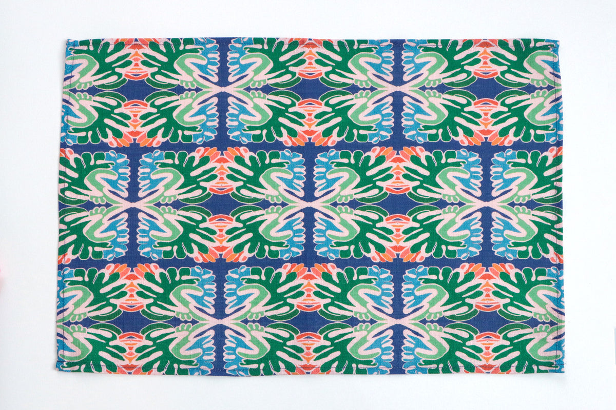 A rectangular fabric placemat with a vibrant, repeating geometric and botanical pattern in green, blue, coral pink, and cream on a white background. Designed by artist Leila Simon Hayes.