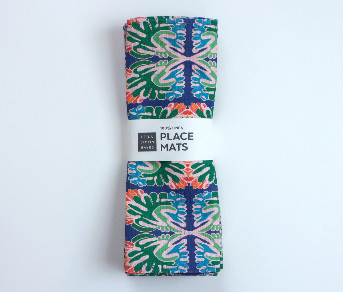 A folded linen place mat with a vibrant, symmetrical mandala-style pattern in green, blue, pink, and orange, wrapped in a white paper band that reads '100% LINEN PLACE MATS' and shows the 'Leila Simon Hayes' brand logo. Designed by artist Leila Simon Hayes.