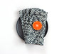 A folded cloth napkin with a black and white floral pattern, styled on a black plate with a bright orange slice on top, set against a plain white background. Designed by artist Leila Simon Hayes.