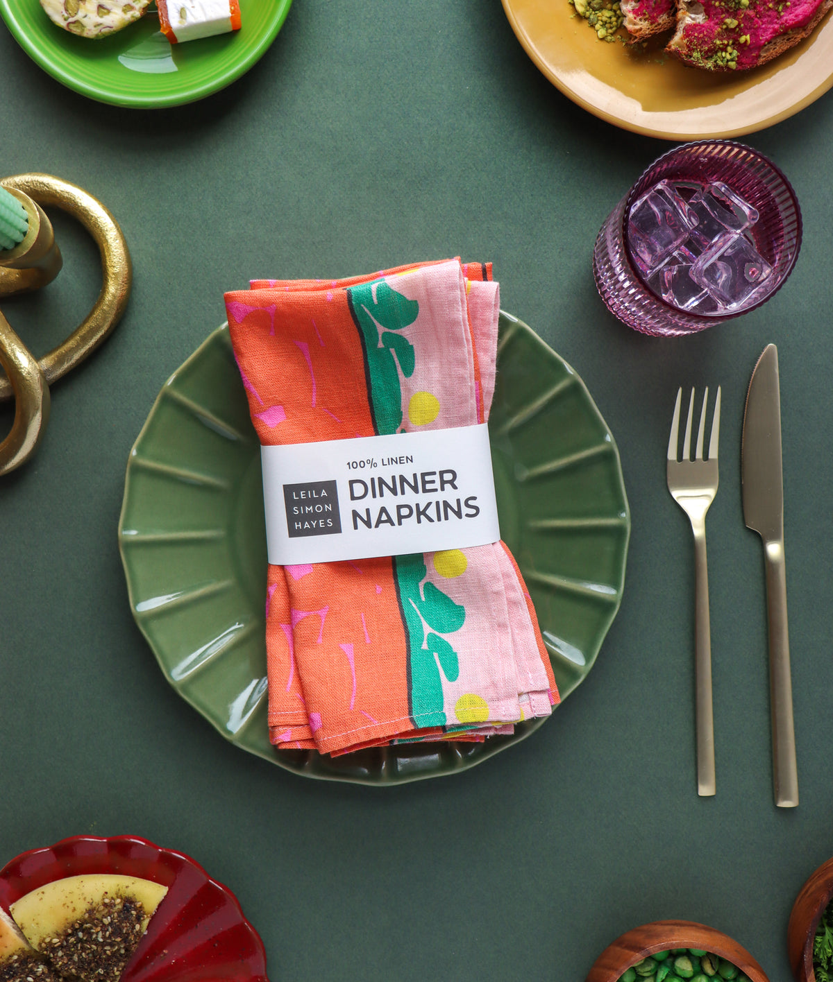 A set of colorful printed cloth napkins wrapped in a paper band, arranged on a green plate on a dark green table with silverware and other dining props. Designed by artist Leila Simon Hayes.
