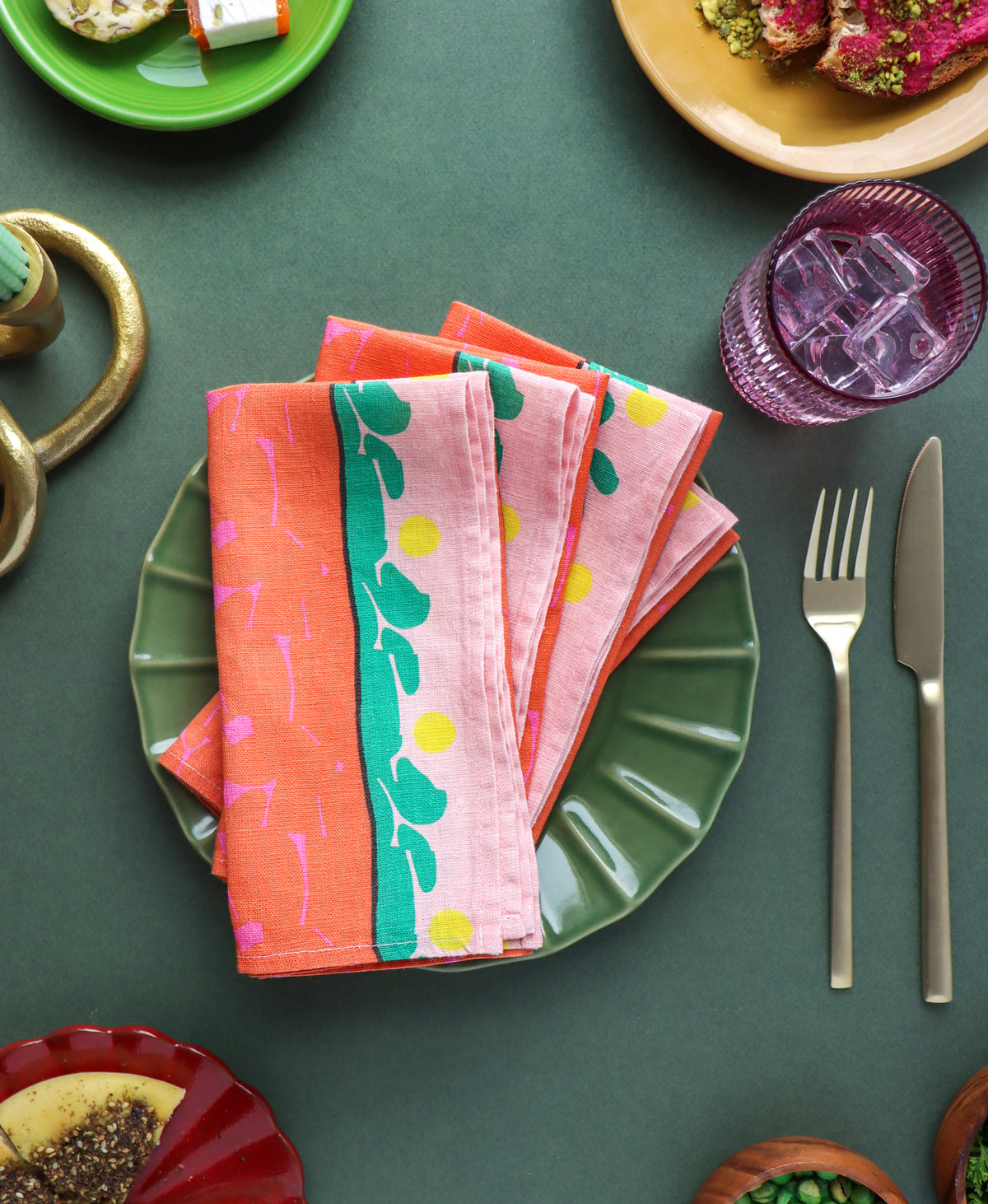 Designed by artist Leila Simon Hayes. A stack of folded patterned linen napkins in coral, pink, and green tones, arranged on a green plate in a styled dining setting with a fork, knife, and other dishes around them.