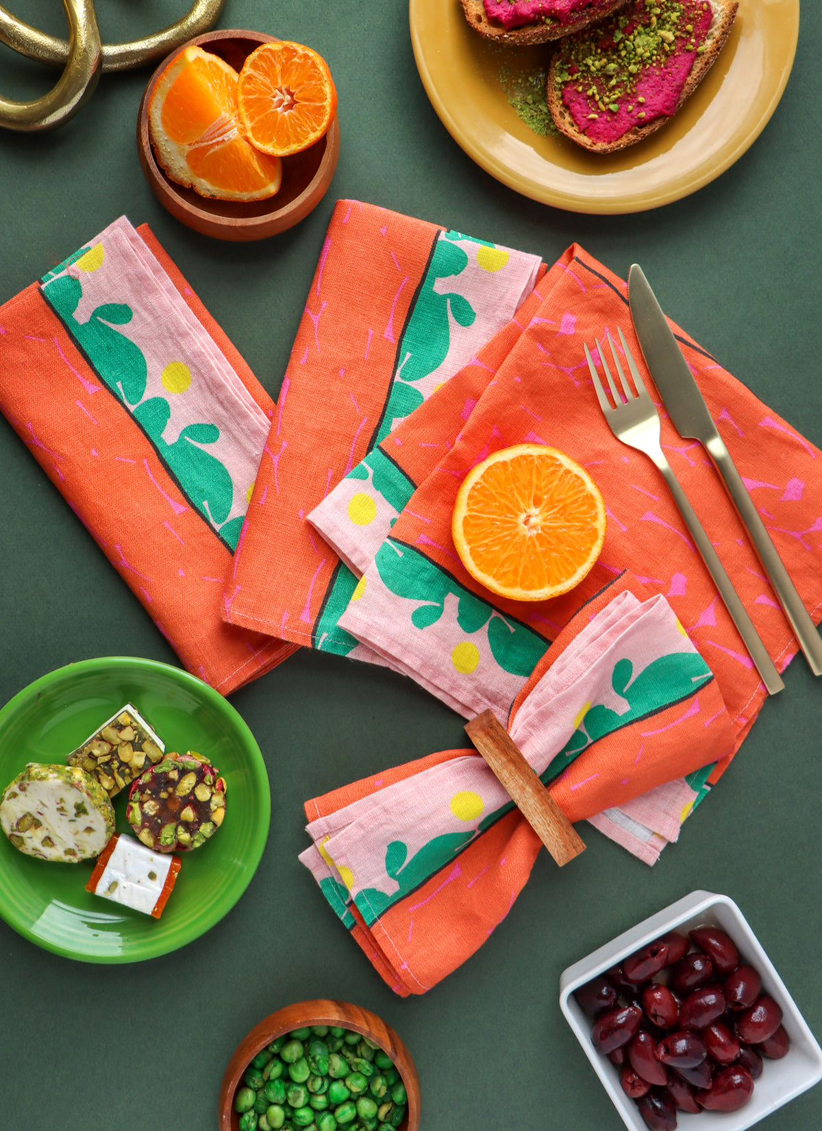 Designed by artist Leila Simon Hayes. A styled flat-lay of folded orange and pink patterned napkins with green leaf motifs, arranged on a dark green tabletop with food props including sliced oranges, toast, green beans, pistachios, and cherries.