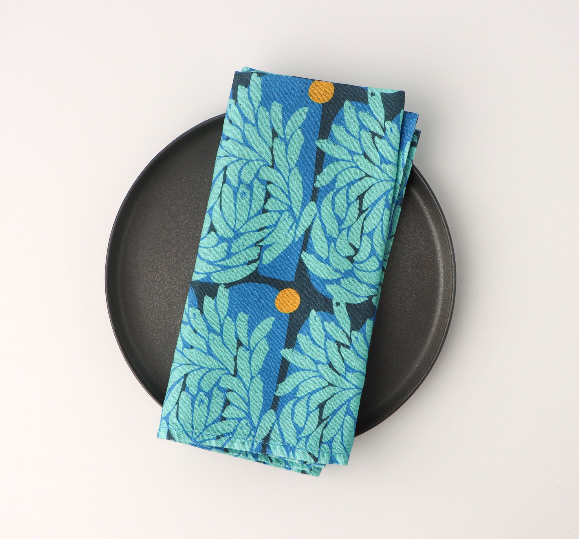 Blue floral leaf-patterned napkin on a black plate against a white background. Designed by artist Leila Simon Hayes.