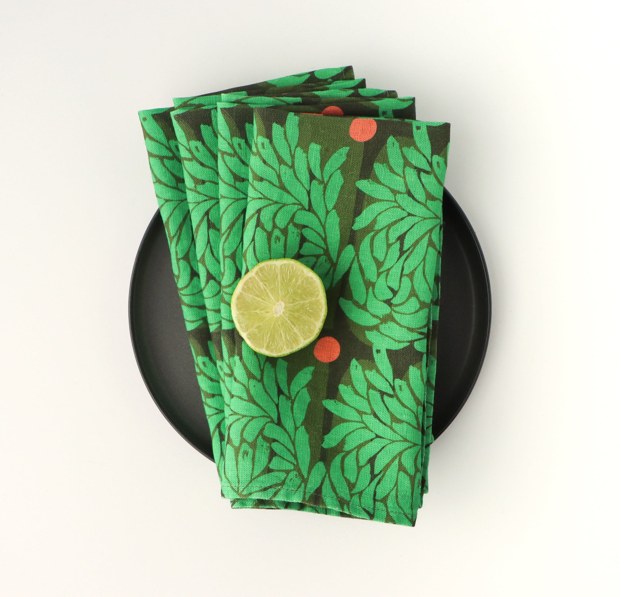 Four green and floral leaf-patterned dinner napkins folded and layered over a black plate, designed by artist Leila Simon Hayes.