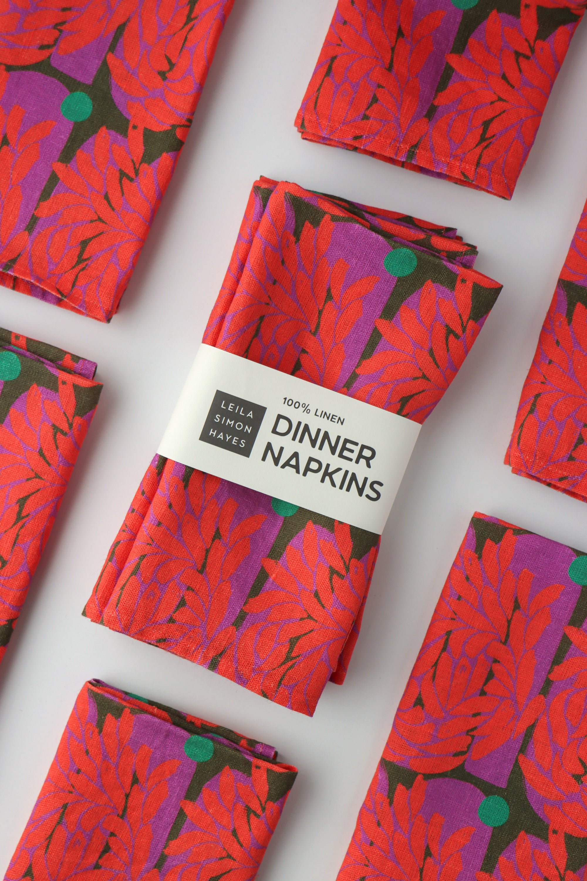 Pack of four packaged dinner napkins with a vibrant red floral pattern with a label saying "100% Linen Dinner Napkins", designed by artist Leila Simon Hayes.