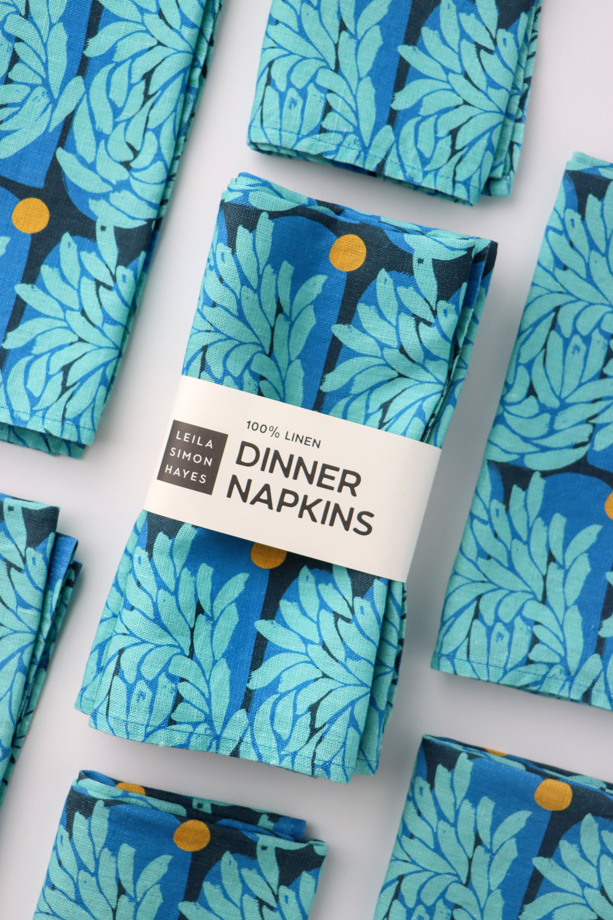 Pack of four,  bright blue floral patterned dinner napkins with a label saying "100% Linen Dinner Napkins", designed by artist Leila Simon Hayes.
