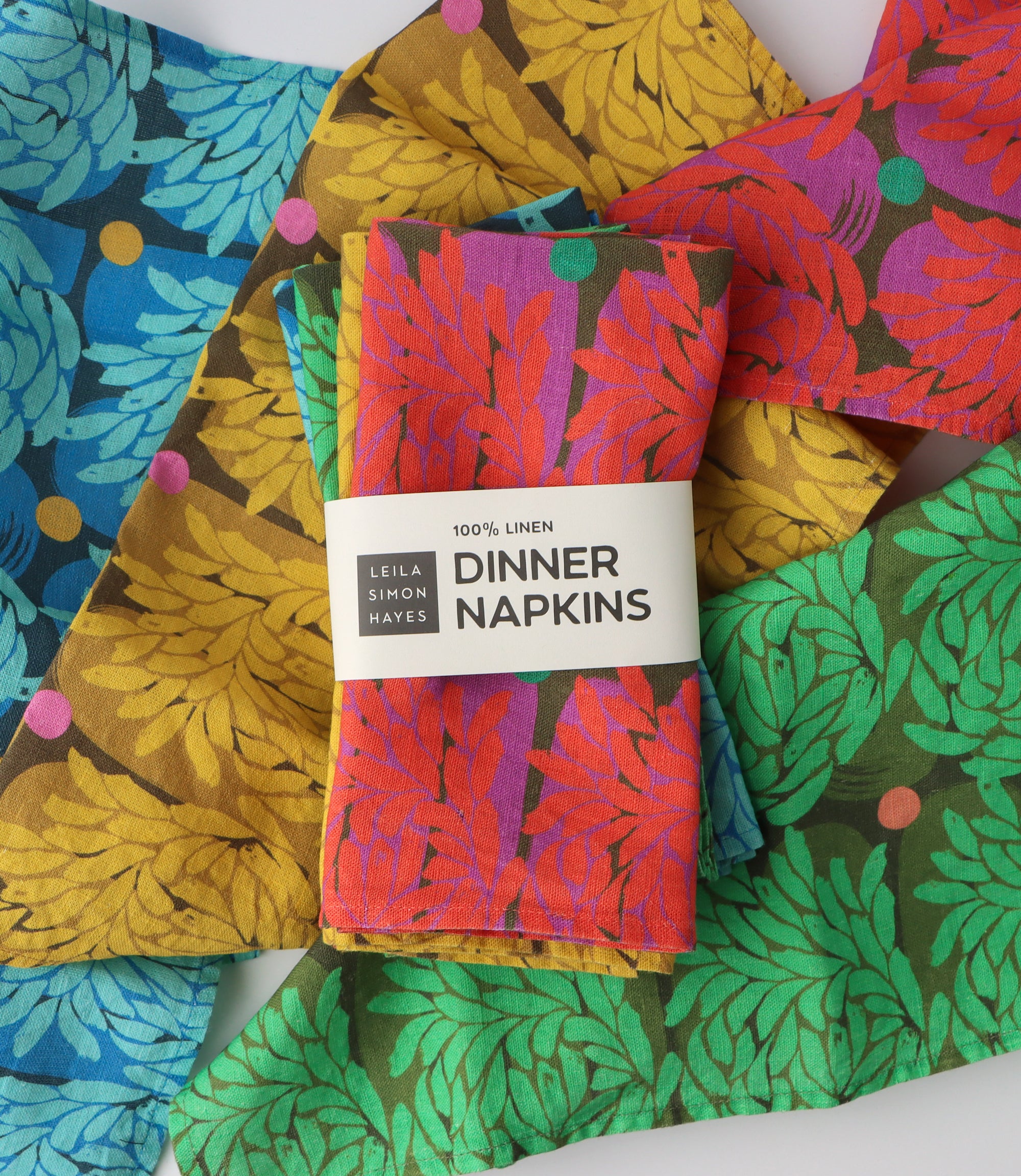 A pack of four colorful dinner napkins with a brand label saying "100% Linen Dinner Napkins" designed by artist Leila Simon Hayes. The background is napkins with the same design layered behind.