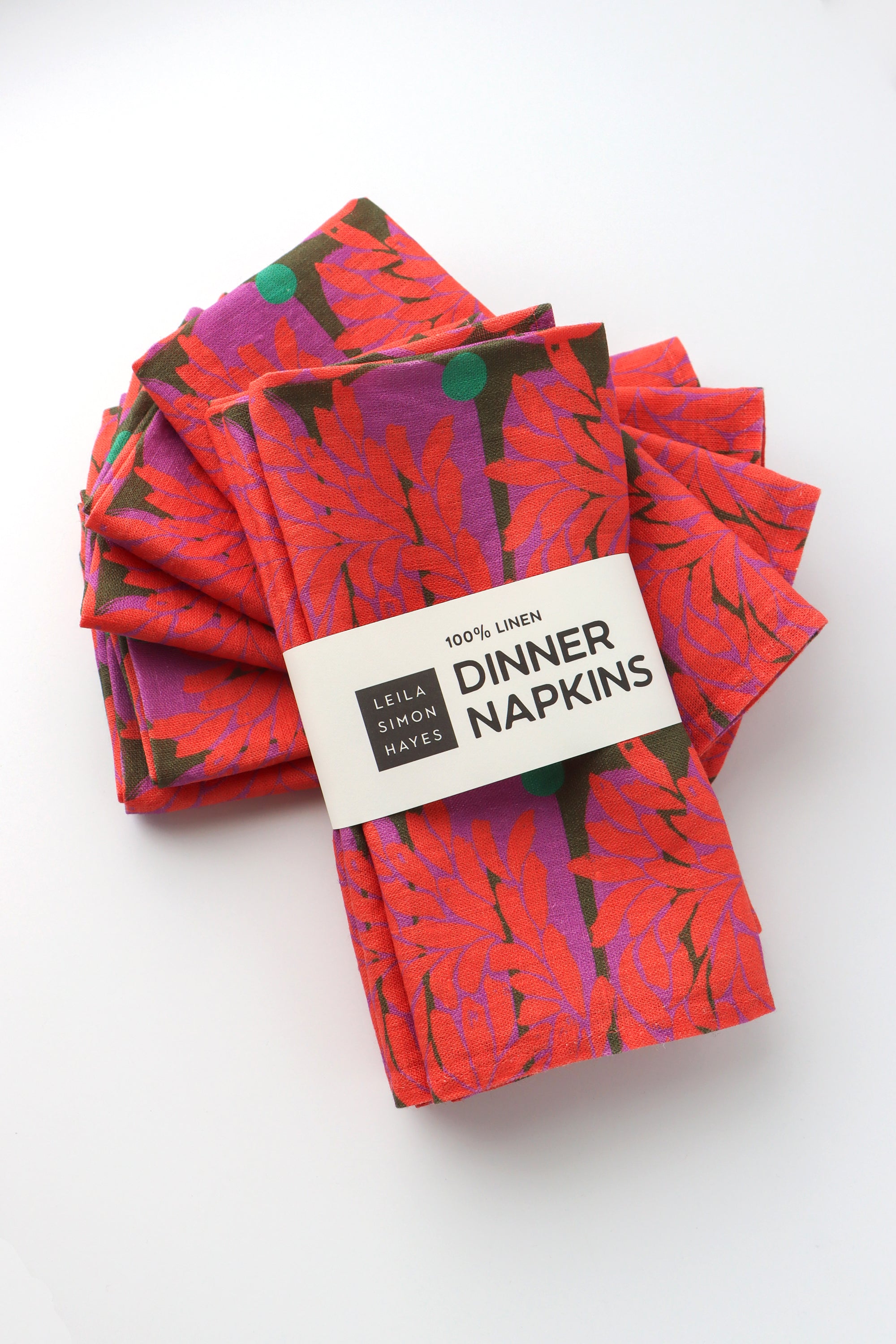 Pack of four packaged dinner napkins with a vibrant red floral pattern with a label saying "100% Linen Dinner Napkins", designed by artist Leila Simon Hayes.