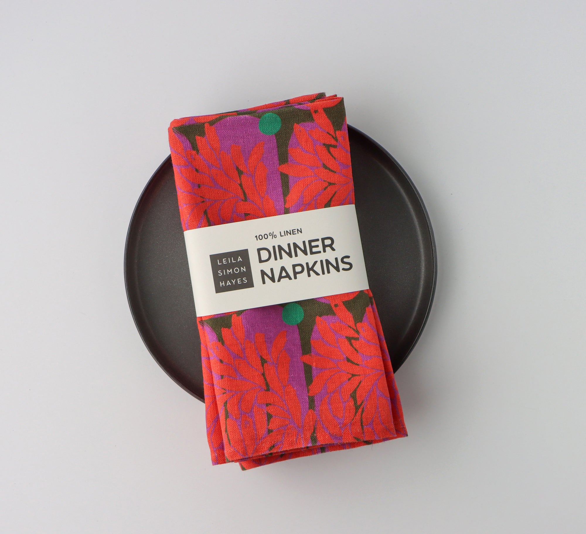 Pack of four packaged dinner napkins with a vibrant red floral pattern with a label saying "100% Linen Dinner Napkins", designed by artist Leila Simon Hayes.