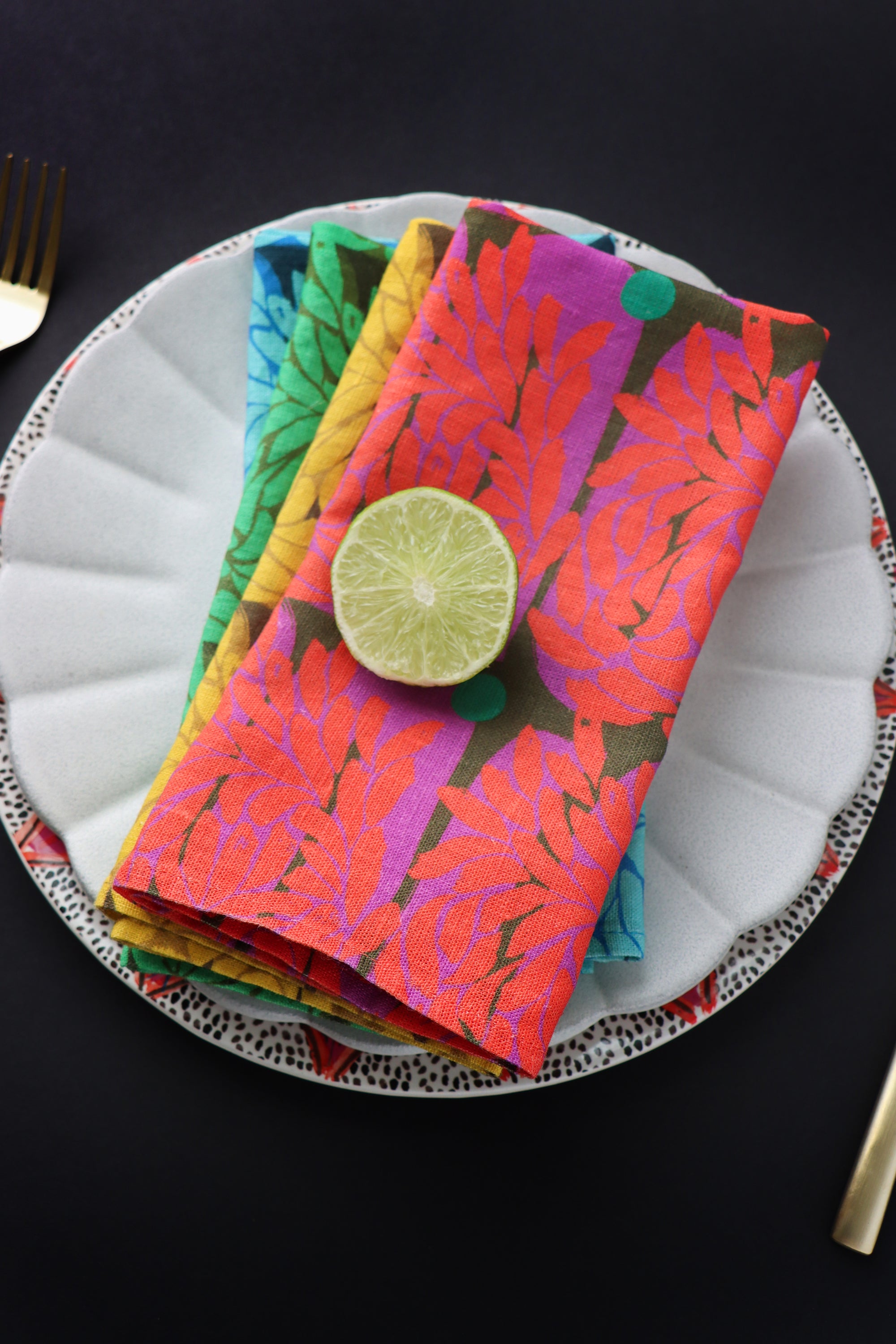 A pack of four colorful, floral napkins on a plate with a lime wedge on a dark background. Designed by artist Leila Simon Hayes.