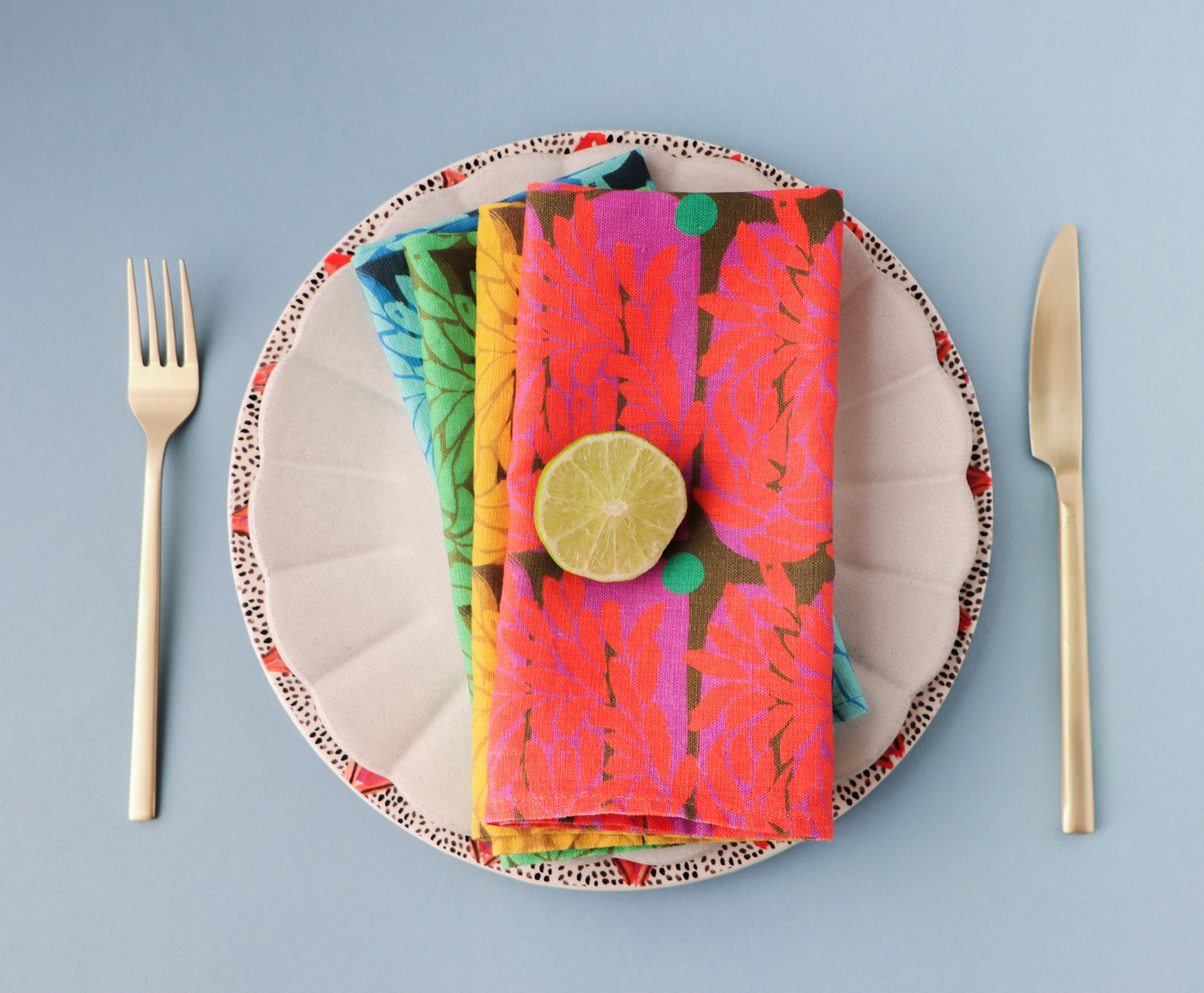 Four colorful, floral napkins layered on a plate with cutlery against a blue background. Designed by artist Leila Simon Hayes.