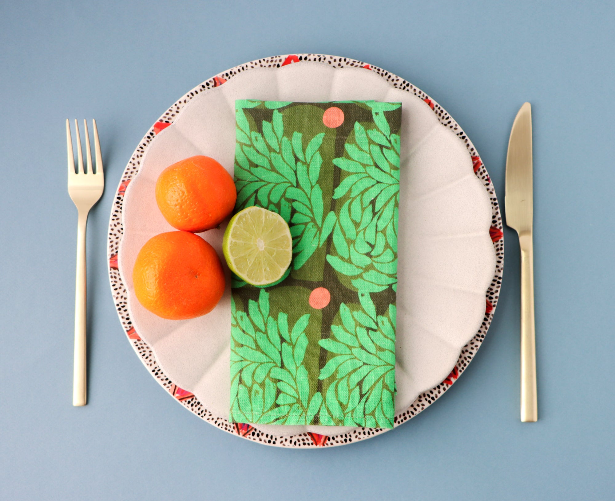 Plate with a green leafy patterned napkin, oranges, and a lime on a blue background. Designed by artist Leila Simon Hayes.