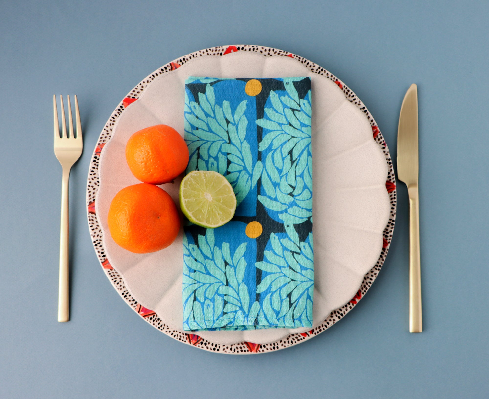 Plate with a blue floral napkin, oranges, and a lime on a blue background. The dinner napkin is designed by artist Leila Simon Hayes.