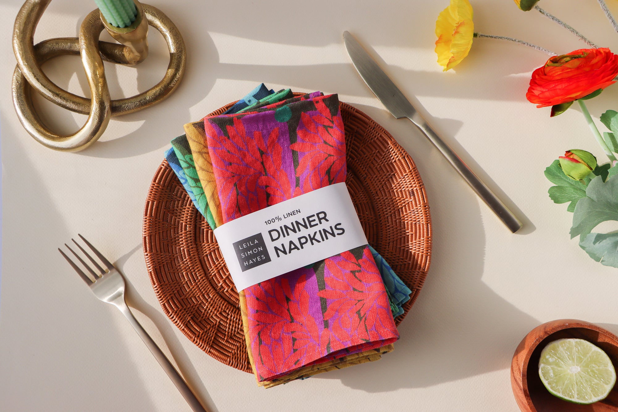 A pack of four colorful, aesthetic, linen dinner napkins on a woven plate with decorative elements.A label is around the napkins saying "100% Linen Dinner Napkins", designed by artist Leila Simon Hayes.