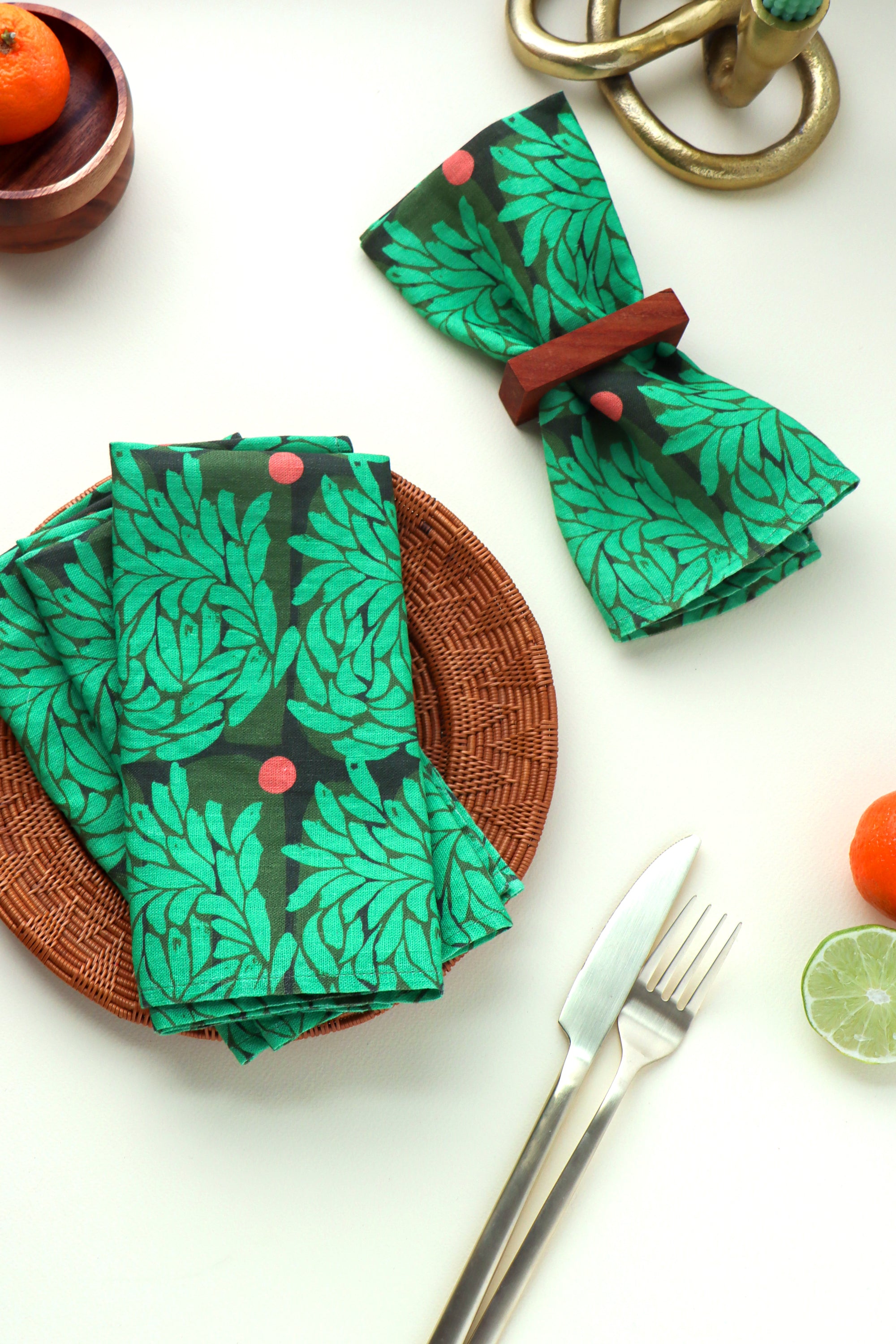 Green abstract floral leaf-patterned napkins on a white surface with cutlery and fruit. Napkins are folded and designed by artist Leila Simon Hayes.