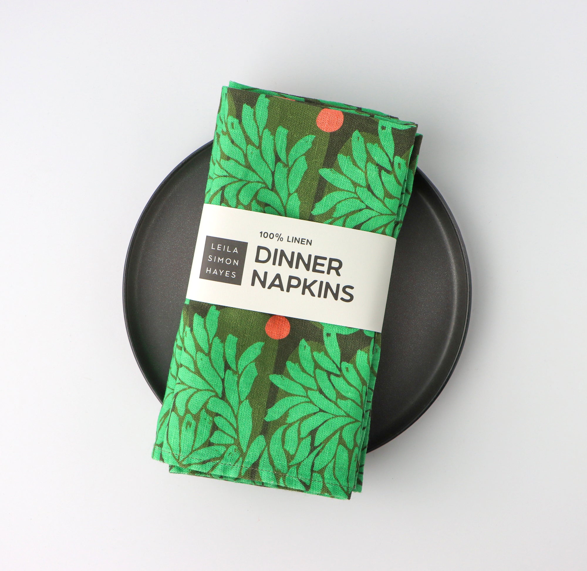 Pack of four, green floral leaf-patterned dinner napkins with a label saying "100% Linen Dinner Napkins", designed by artist Leila Simon Hayes. On a black plate with a white background.