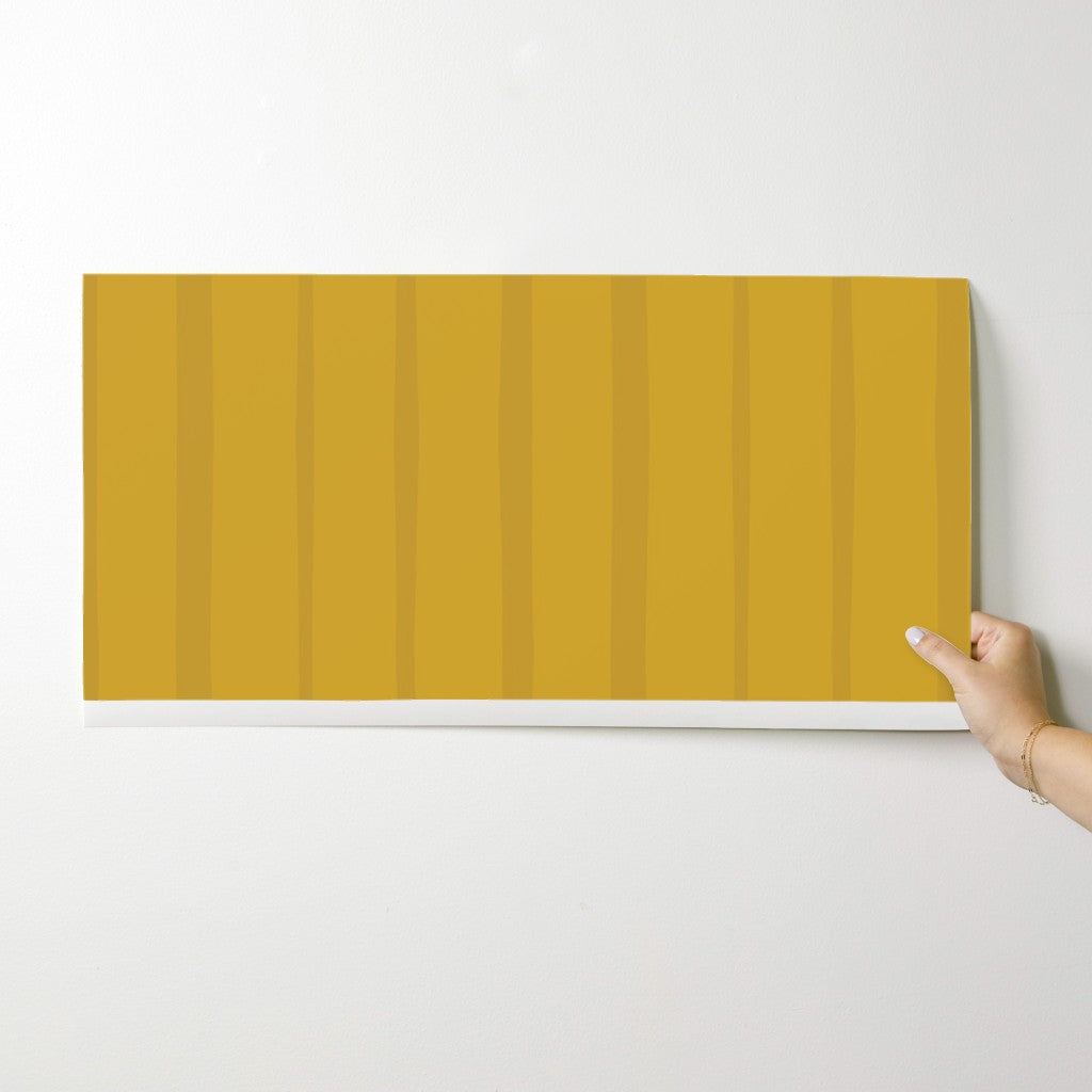 Hand holding a mustard yellow wallpaper swatch with vertical stripes on a white background. Designed by artist Leila Simon Hayes.