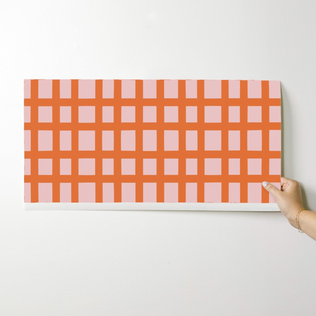 Hand holding a wallpaper swatch with a pink and orange checkered pattern against a white background. Designed by artist Leila Simon Hayes.