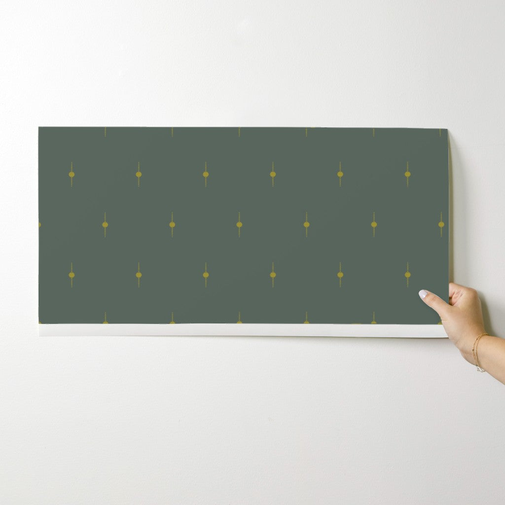 Hand holding an olive green wallpaper swatch with gold geometric star pattern against a white background. Designed by artist Leila Simon Hayes. 