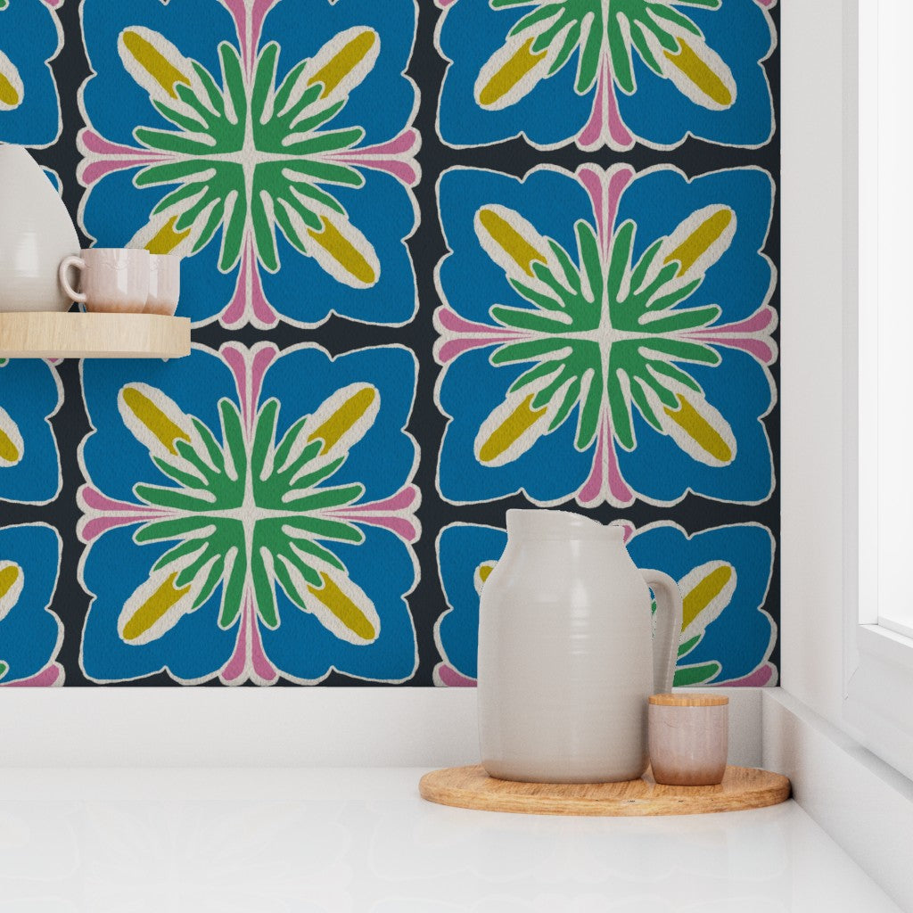 A close-up of a kitchen backsplash featuring bold, colorful floral patterned tiles in blue, green, yellow, pink, and black. A white ceramic pitcher and small cup sit on a wooden coaster on the white countertop below. Designed by artist Leila Simon Hayes.