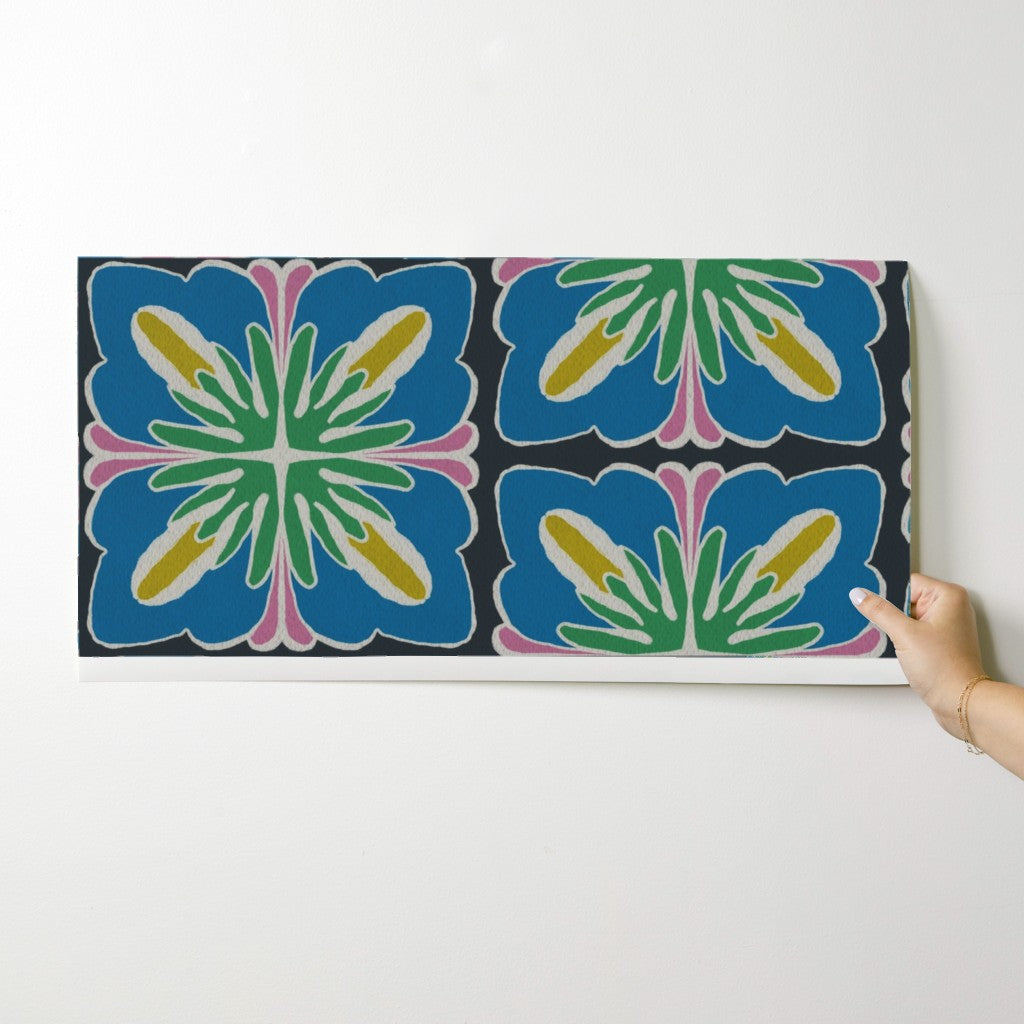A rectangular wall art print or panel with a bold retro floral pattern in blue, green, pink, yellow, and white on a dark background, shown against a light gray wall with a hand holding the right edge. Designed by artist Leila Simon Hayes.