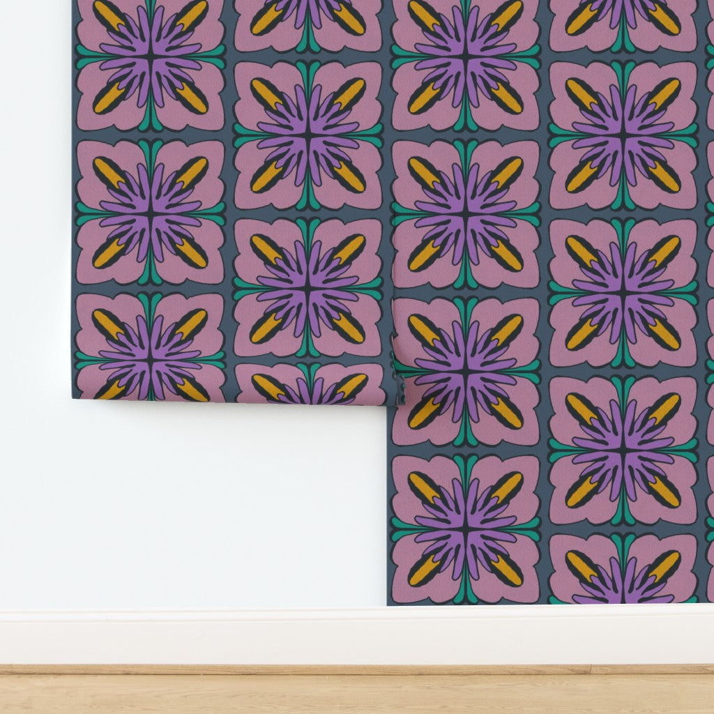 A roll of wallpaper with a retro 1970s floral pattern, partially unrolled on a white wall above a light wood floor. The pattern features purple and lavender flowers with yellow centers and teal accents on a dark gray background. Designed by artist Leila Simon Hayes.