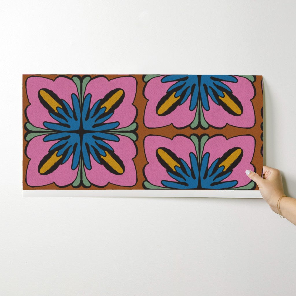 A rectangular wall art print or canvas with a bold retro floral pattern in pink, blue, yellow, green, and brown, shown hanging against a white wall with a hand holding the right edge. Designed by artist Leila Simon Hayes.