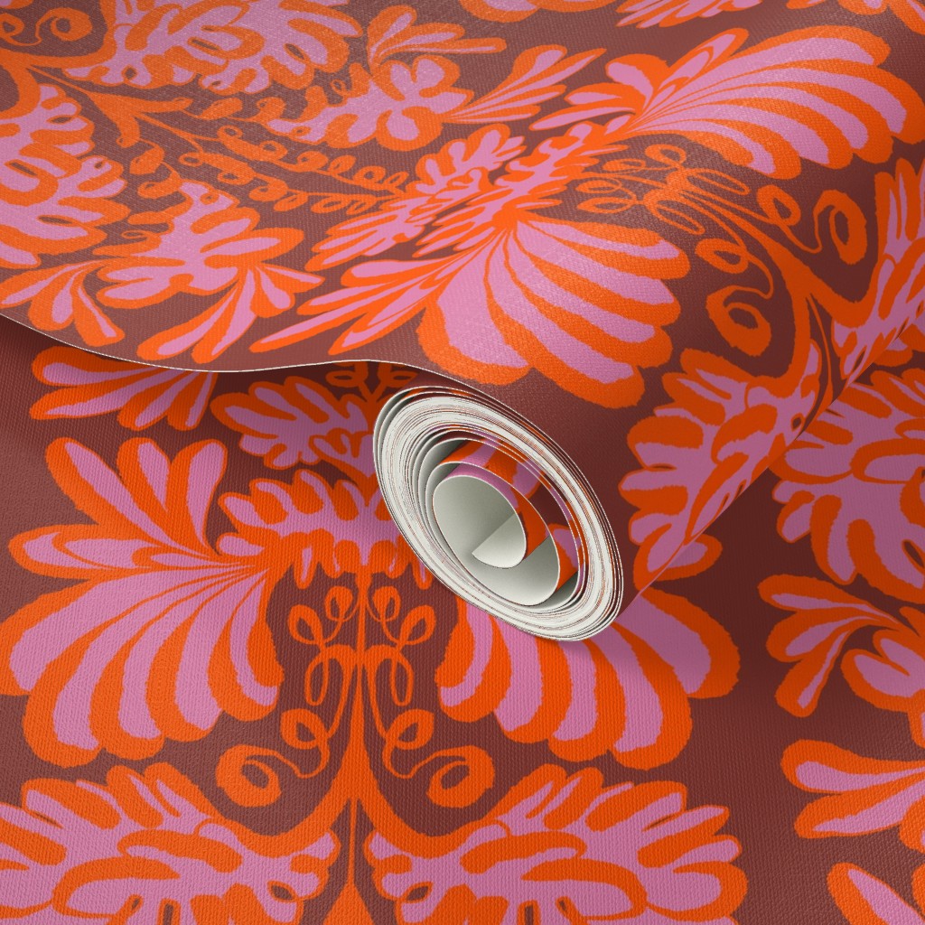 A roll of patterned wallpaper or fabric shown close up, with a rolled silver core visible. The design features bold retro floral and damask motifs in bright orange and light pink on a warm reddish-brown background. Designed by artist Leila Simon Hayes.