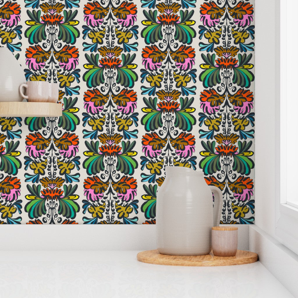 A colorful floral wallpaper with a vertical damask pattern in pink, orange, green, yellow, and blue on a white background, shown in a styled room setting with a white pitcher and cup on a wooden tray. Designed by artist Leila Simon Hayes.