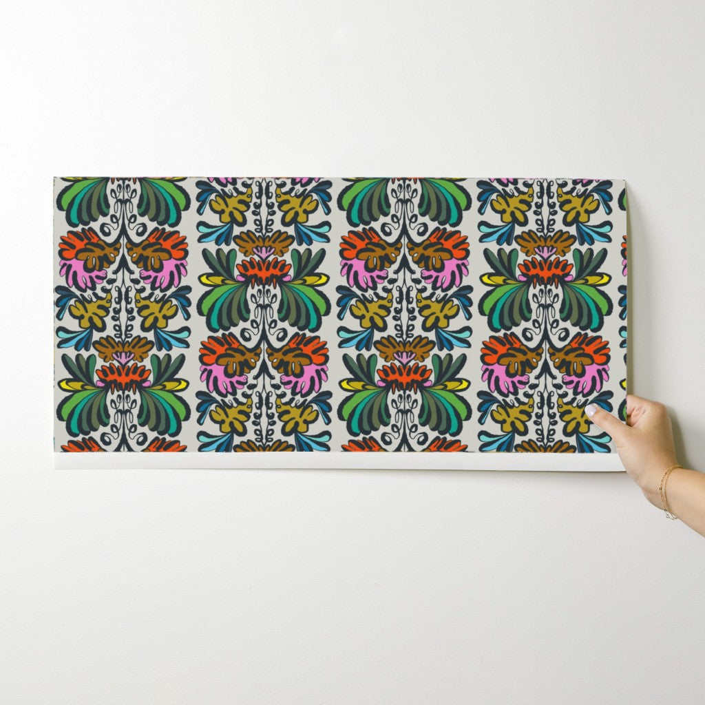 A rectangular canvas print featuring a vibrant, symmetrical floral and ornamental pattern in multiple colors, shown hanging on a plain white wall with a hand holding the right edge. Designed by artist Leila Simon Hayes.