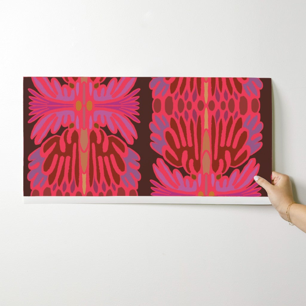 A rectangular wall art print with a bold abstract floral pattern in pink, magenta, purple, and brown, shown hanging on a light gray wall with a hand holding the bottom right corner. Designed by artist Leila Simon Hayes.