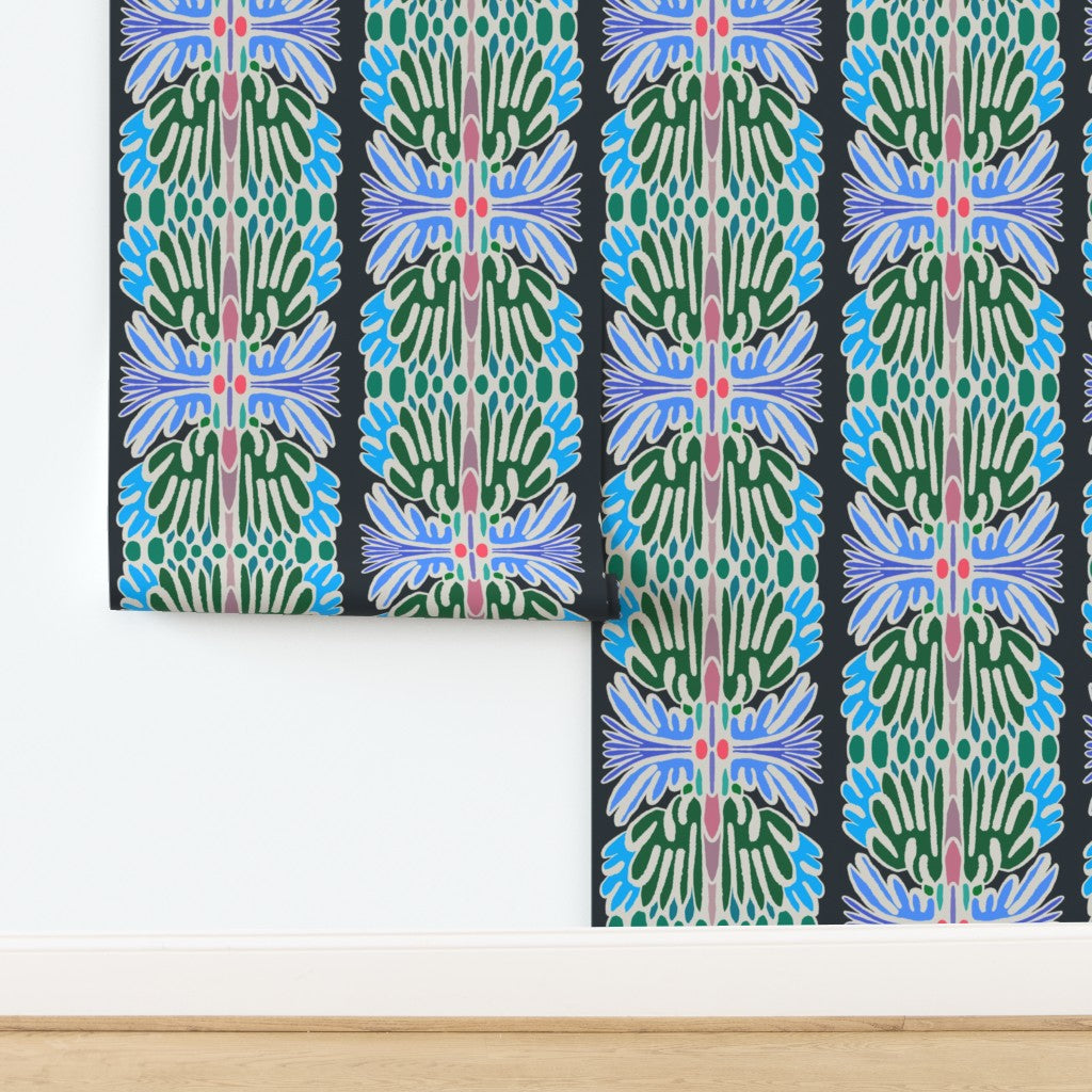 A folded and partially unrolled sheet of wallpaper with a bold, symmetrical botanical pattern in green, blue, pink, and white on a black background, displayed on a white wall above a light wood floor. Designed by artist Leila Simon Hayes.