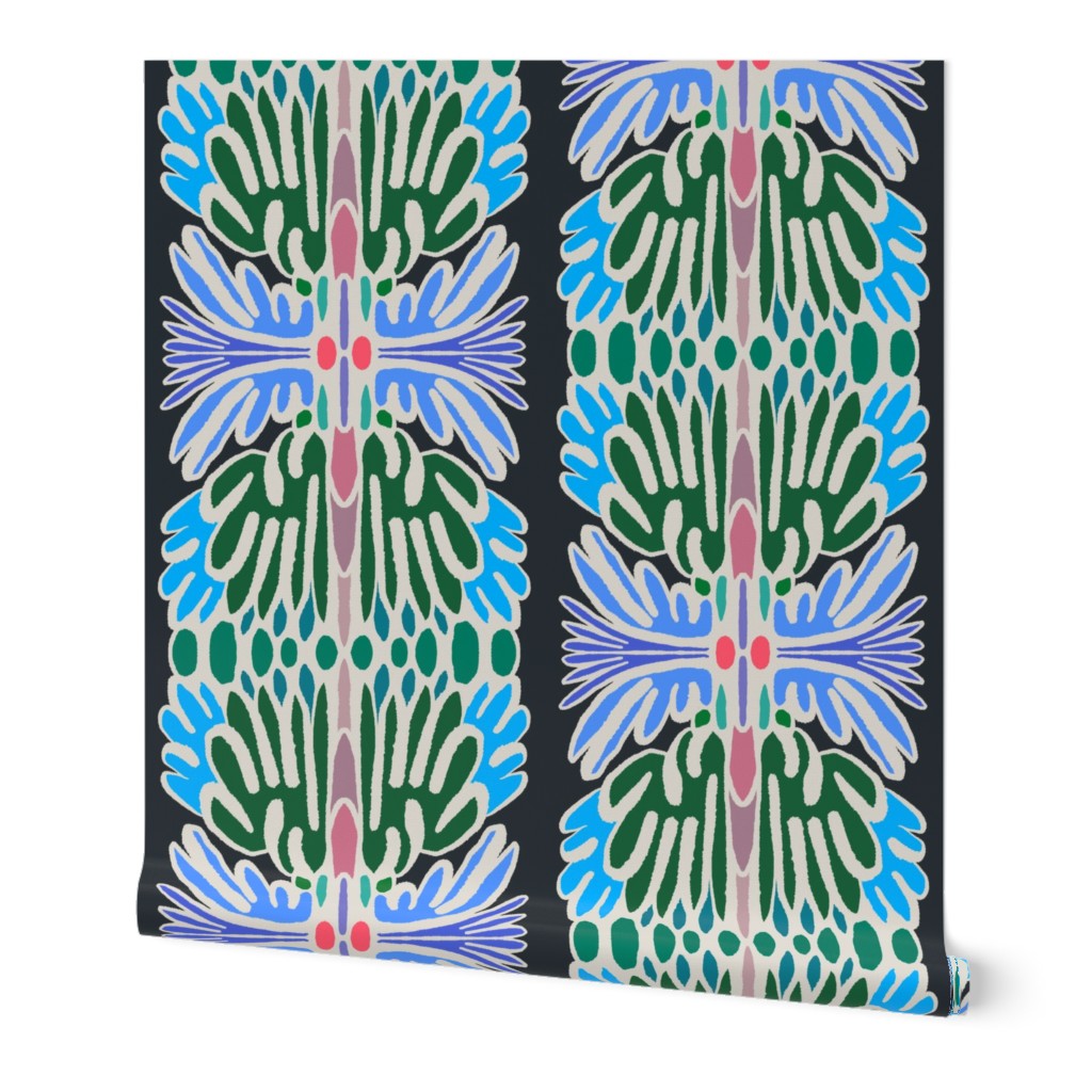 A wallpaper roll with geometric and floral-inspired pattern in green, blue, lavender, cream, and small pink accents on a dark background, shown against a plain white studio backdrop. Designed by artist Leila Simon Hayes.
