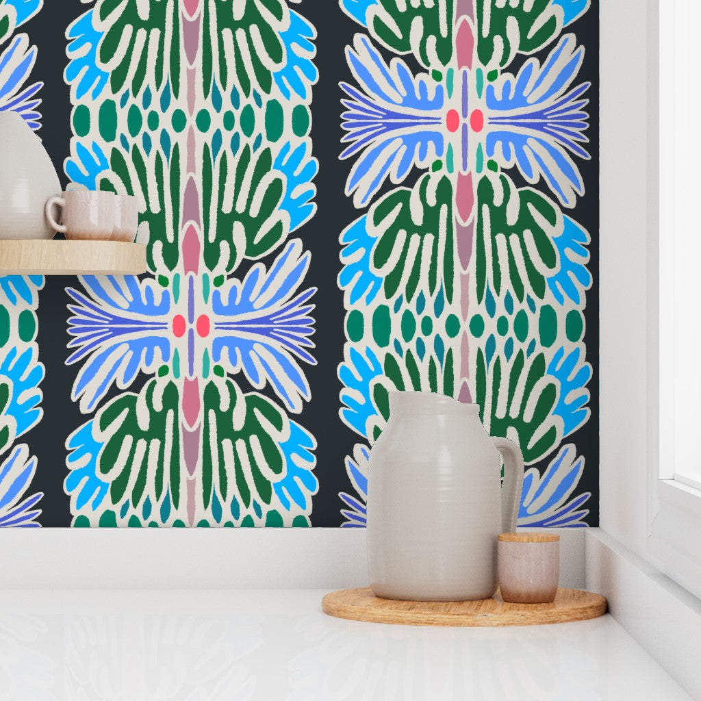 A styled interior corner showing a bold vertical patterned wallpaper with green, blue, white, and pink motifs on a black background, mounted on a white wall above a white surface with a ceramic pitcher and small cup on a wooden tray. Designed by artist Leila Simon Hayes.
