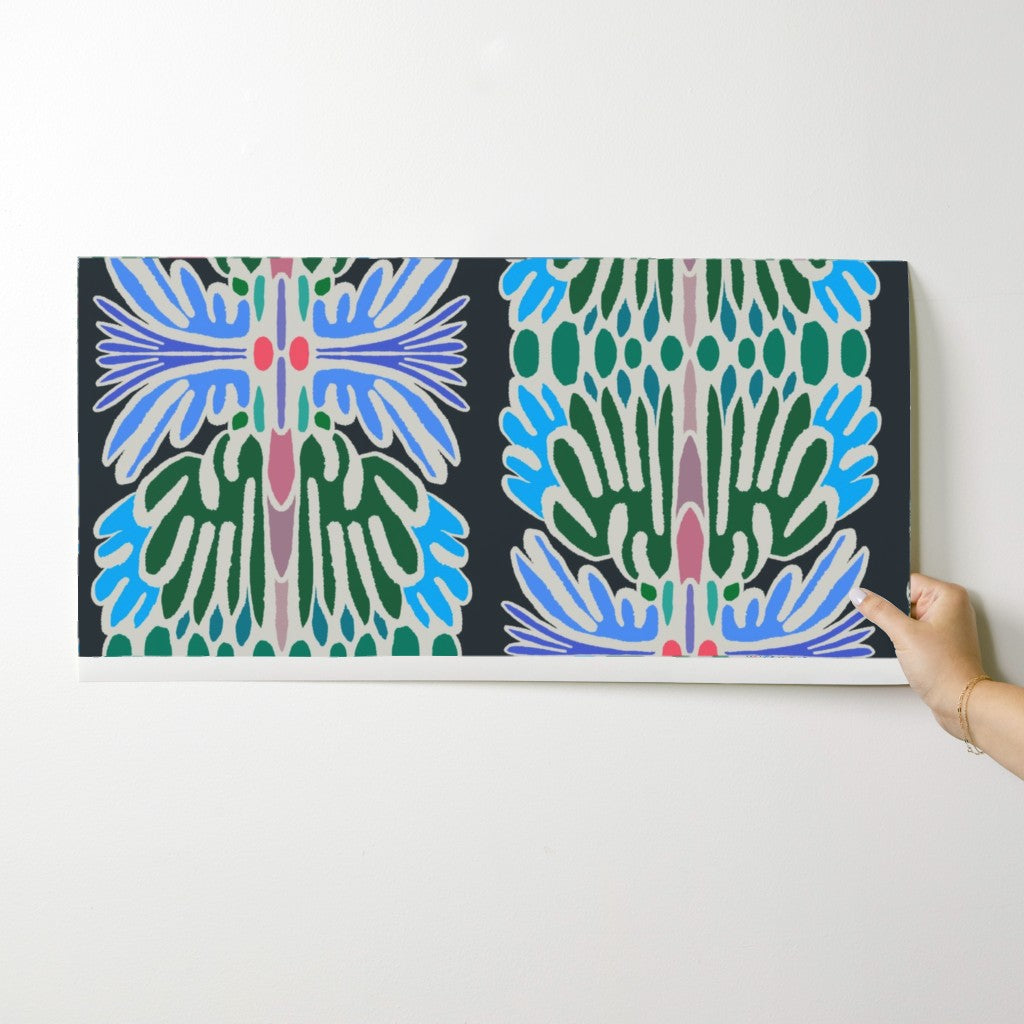 A rectangular canvas print featuring a symmetrical, stylized botanical pattern in blue, green, pink, and white on a black background, shown hanging on a light gray wall with a hand holding the right edge. Designed by artist Leila Simon Hayes.
