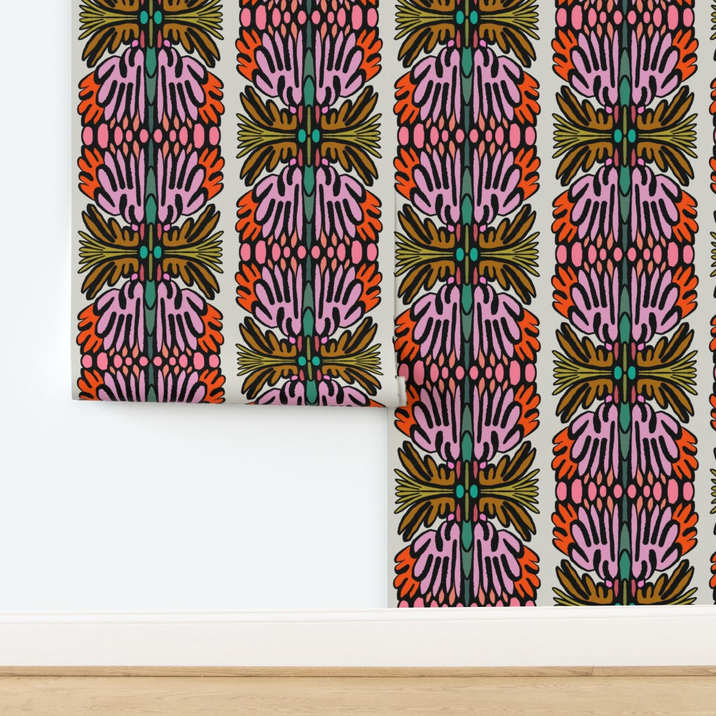 A close-up of wallpaper with a bold vertical pattern featuring pink, orange, olive green, and teal motifs on a white background, shown installed on a wall above a light wood floor. Designed by artist Leila Simon Hayes.