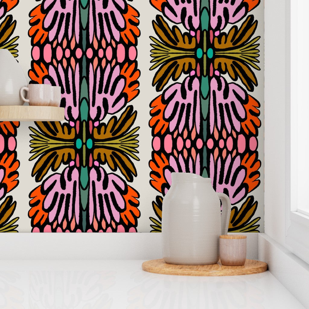 A brightly patterned wallpaper or wall covering with vertical abstract floral motifs in pink, orange, olive green, teal, and black on a white background, shown in a styled interior corner with a white pitcher and small cup on a wooden tray. Designed by artist Leila Simon Hayes.