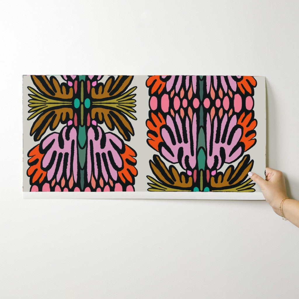 A rectangular wall art print with a bold symmetrical abstract floral pattern in pink, orange, brown, and teal on a white background, being held against a white wall by a person's hand. Designed by artist Leila Simon Hayes.