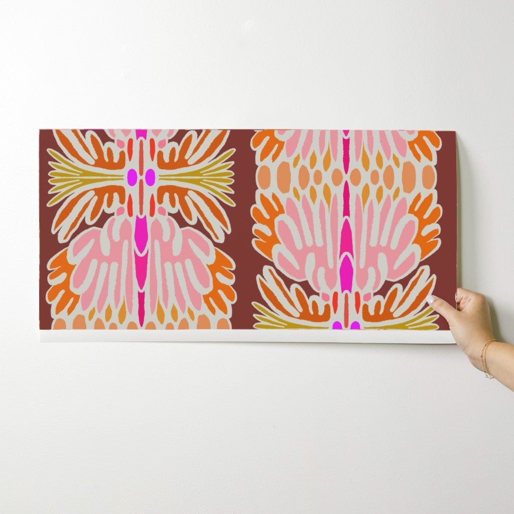 A rectangular wall art print with a symmetrical retro floral pattern in pink, orange, yellow, and magenta on a dark brown background, shown hanging on a light gray wall with a hand holding the right edge. Designed by artist Leila Simon Hayes.