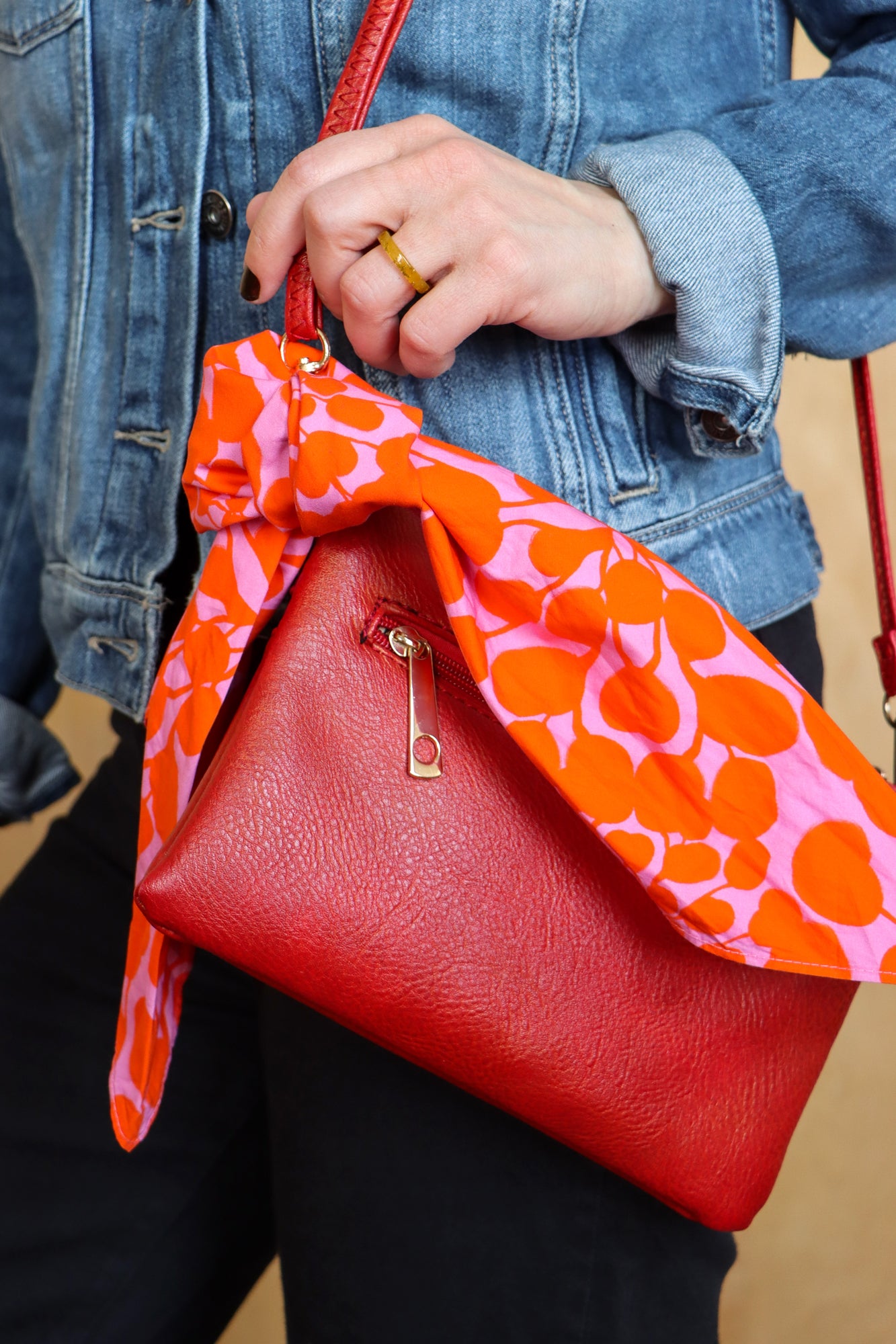 Red handbag with an orange and pink patterned strap held by a person wearing a denim jacket. Designed by artist Leila Simon Hayes.